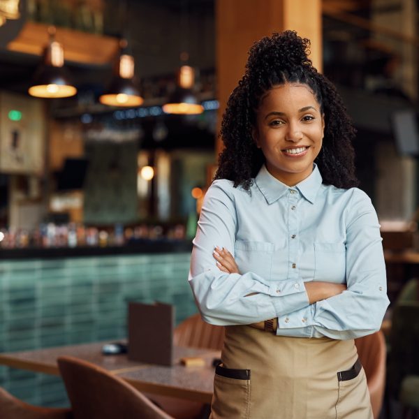 Happy African American waitress with crossed arms in a cafe.,